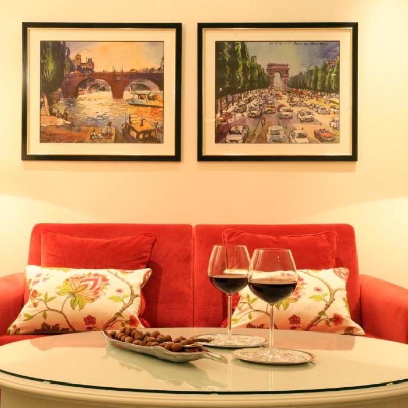 Rotes Sofa, davor ein runder Couchtisch mit 2 Gläsern Rotwein. Im Hintergrund hängen zwei schöne Bilder an der Wand